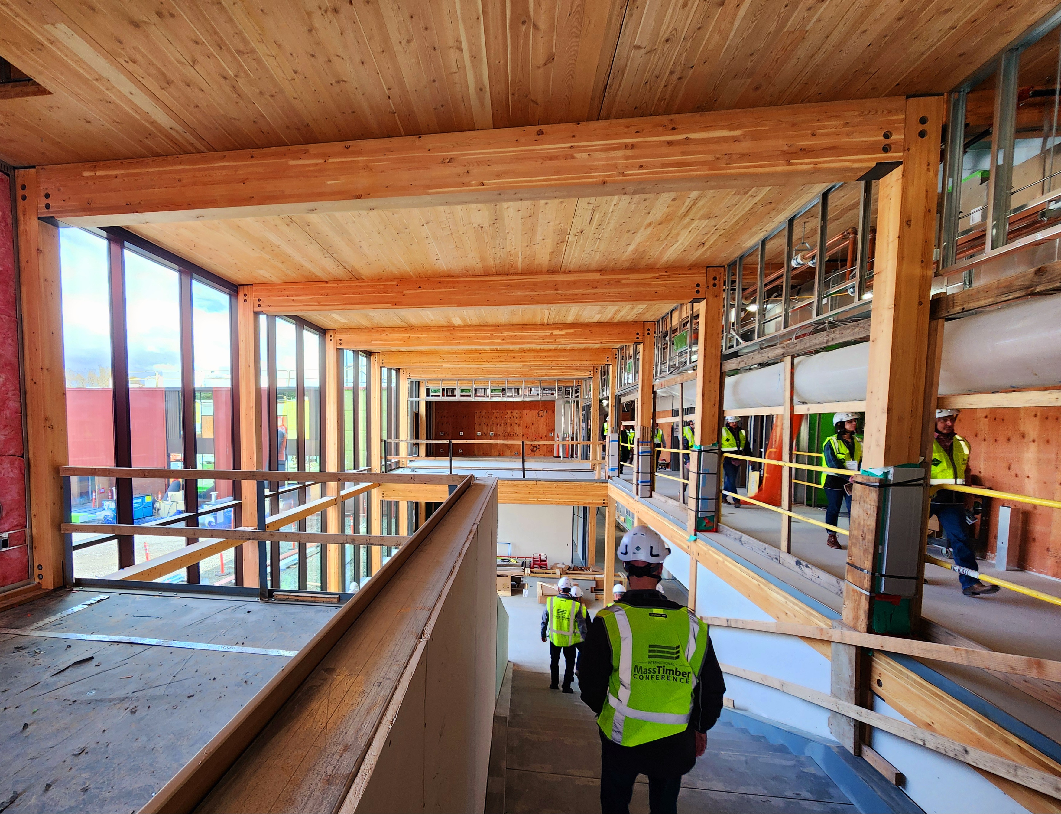 Exposed mass timber with warm colors and people walking down the staircase that is under construction during an International Mass Timber Conference Tour. 