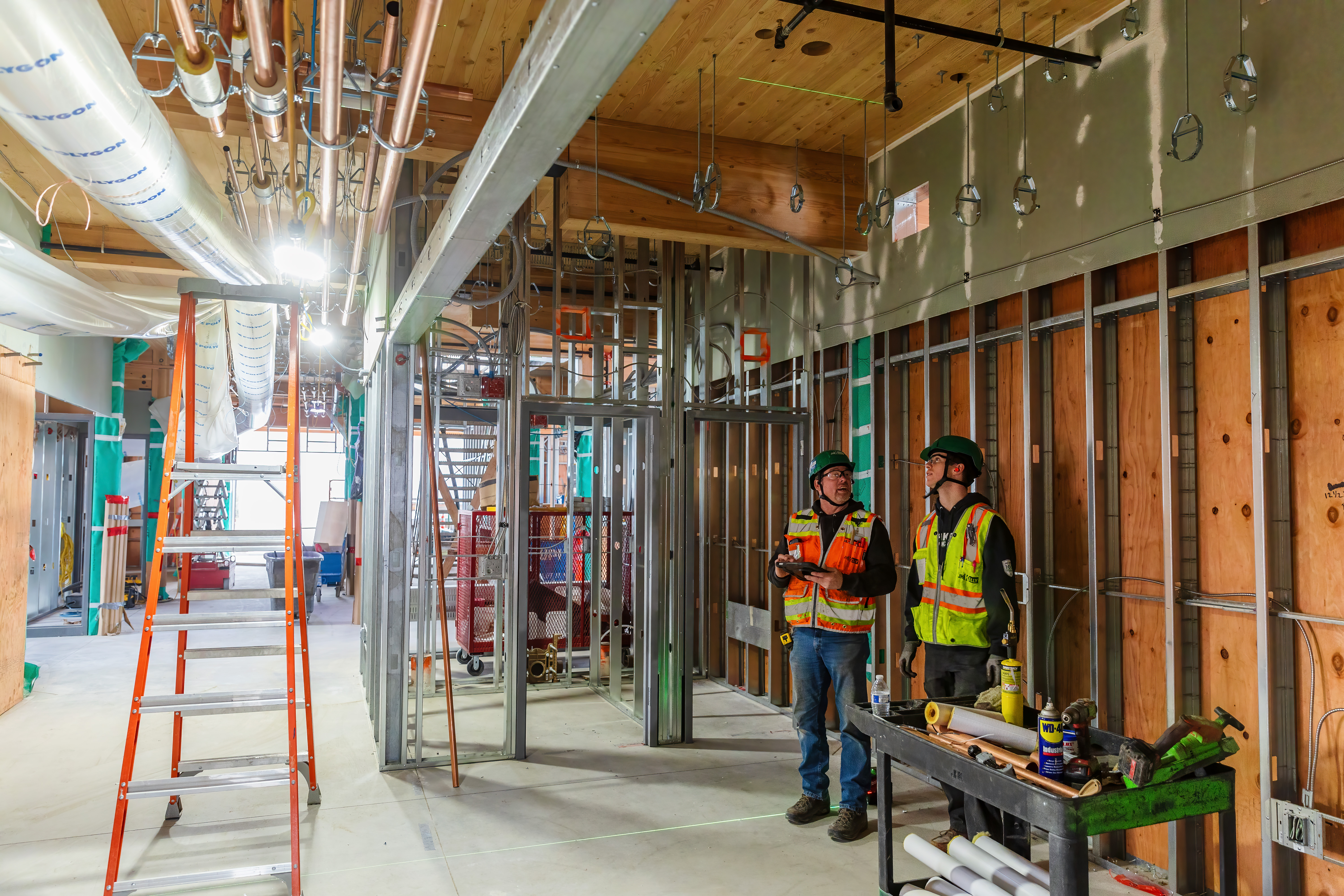 MEP installing with 2 men standing on the right with PPE on like helmets, a vest, jeans. A ladder on the left near open duct work and some exposed mass timber ceiling in the frame. 
