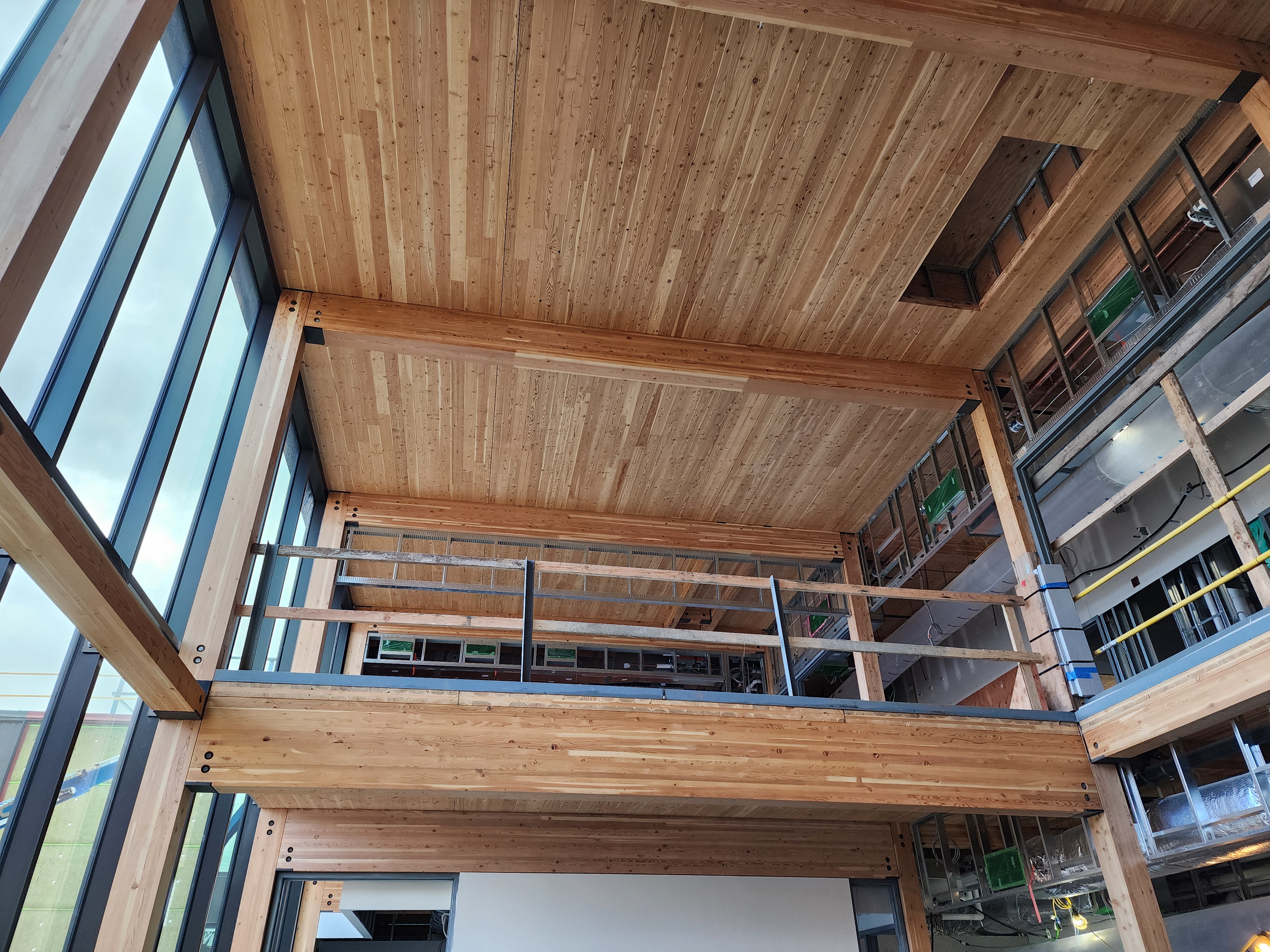 Looking into a mass timber ceiling with panels placed still under construction of an entry way in the building. A landing and hallways are around the outside of the frame. 