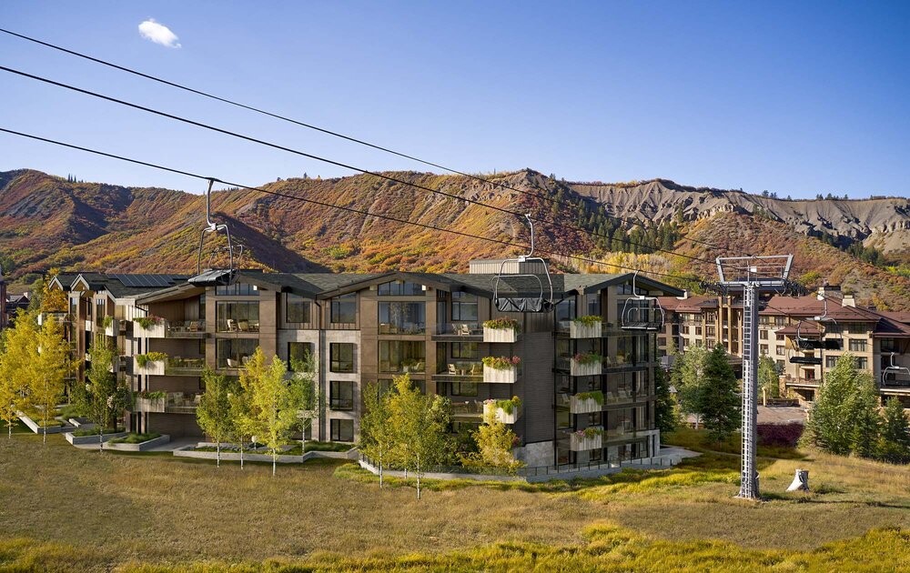The outside of the mass timber Aura Snowmass Village in Colorado Rocky Mountains with ski lifts and green, orange, and yellow deciduous trees during Fall time.
