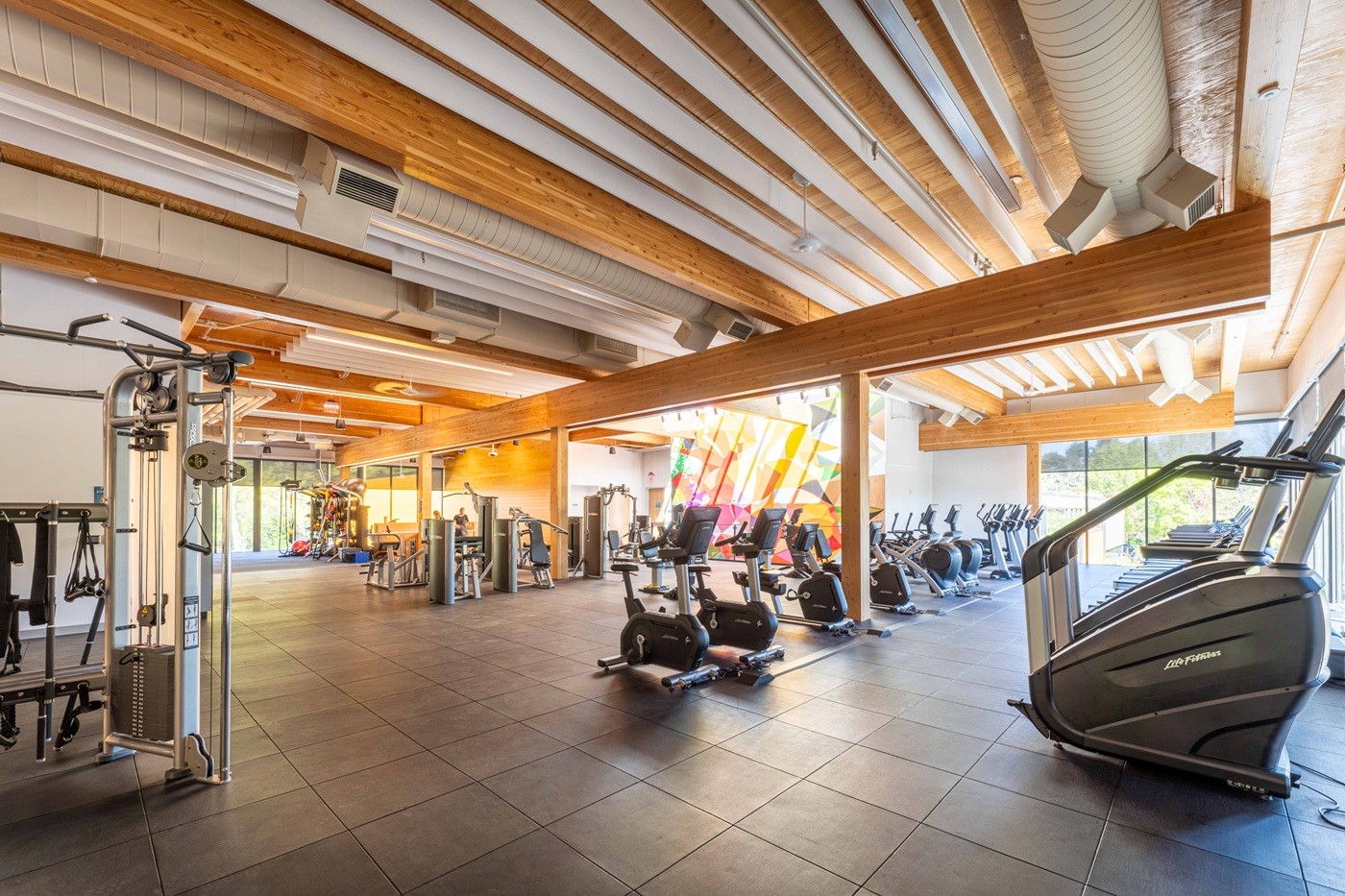 A wide-open fitness center with mass timber beams and columns exposed inside so that it feels warm and welcoming. Fitness equipment is lined up and black acoustic mat is on the top of the floor for sound dampening.