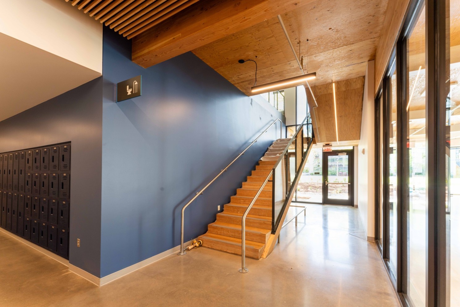 A blue wall inside a hallway corner with lockers and a mass timber staircase with wooden ceiling panels and floor to ceiling windows.