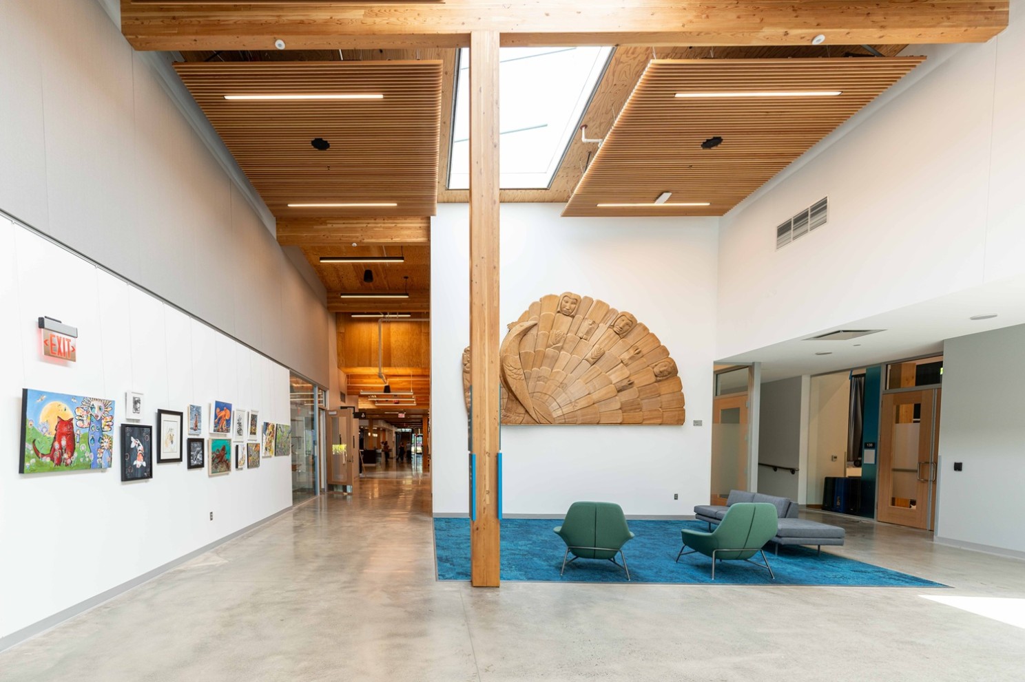 A lobby area with a wooden masterpiece on the wall of tribal origin. A beam made of mass timber in the middle. Ceilings have hanging ascent wood. Artwork is down the hallways with white walls.