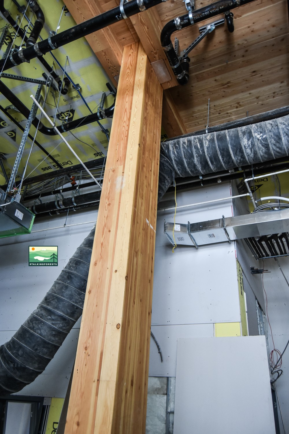 A mass timber beam in the front lobby entry way with duct work still exposed during the construction of the inside of the building. Exposed CLT ceilings and pipe work is seen too.
