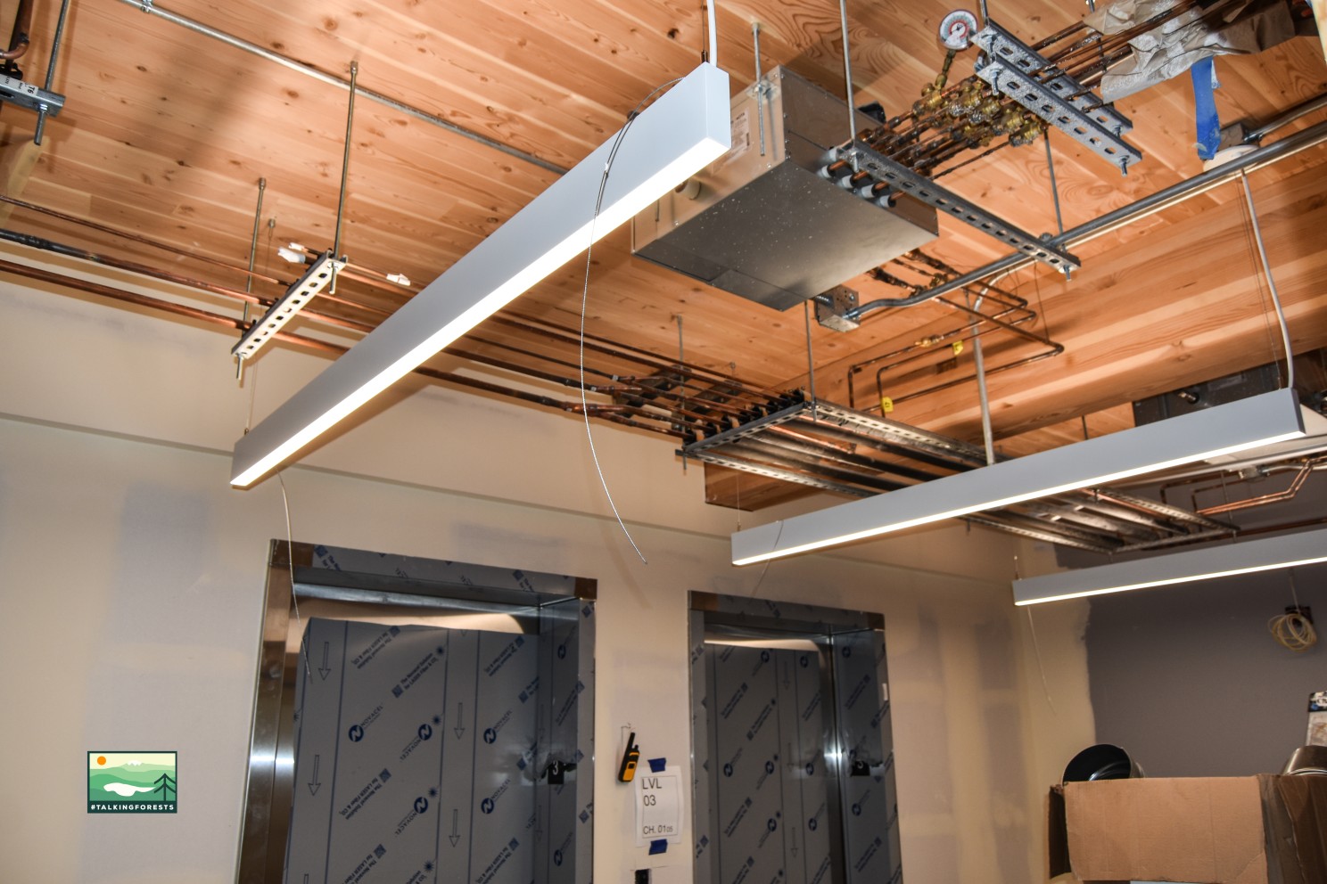 Exposed MEP by the elevator doors that are silver and the building has exposed wood on the ceiling made of mass timber.