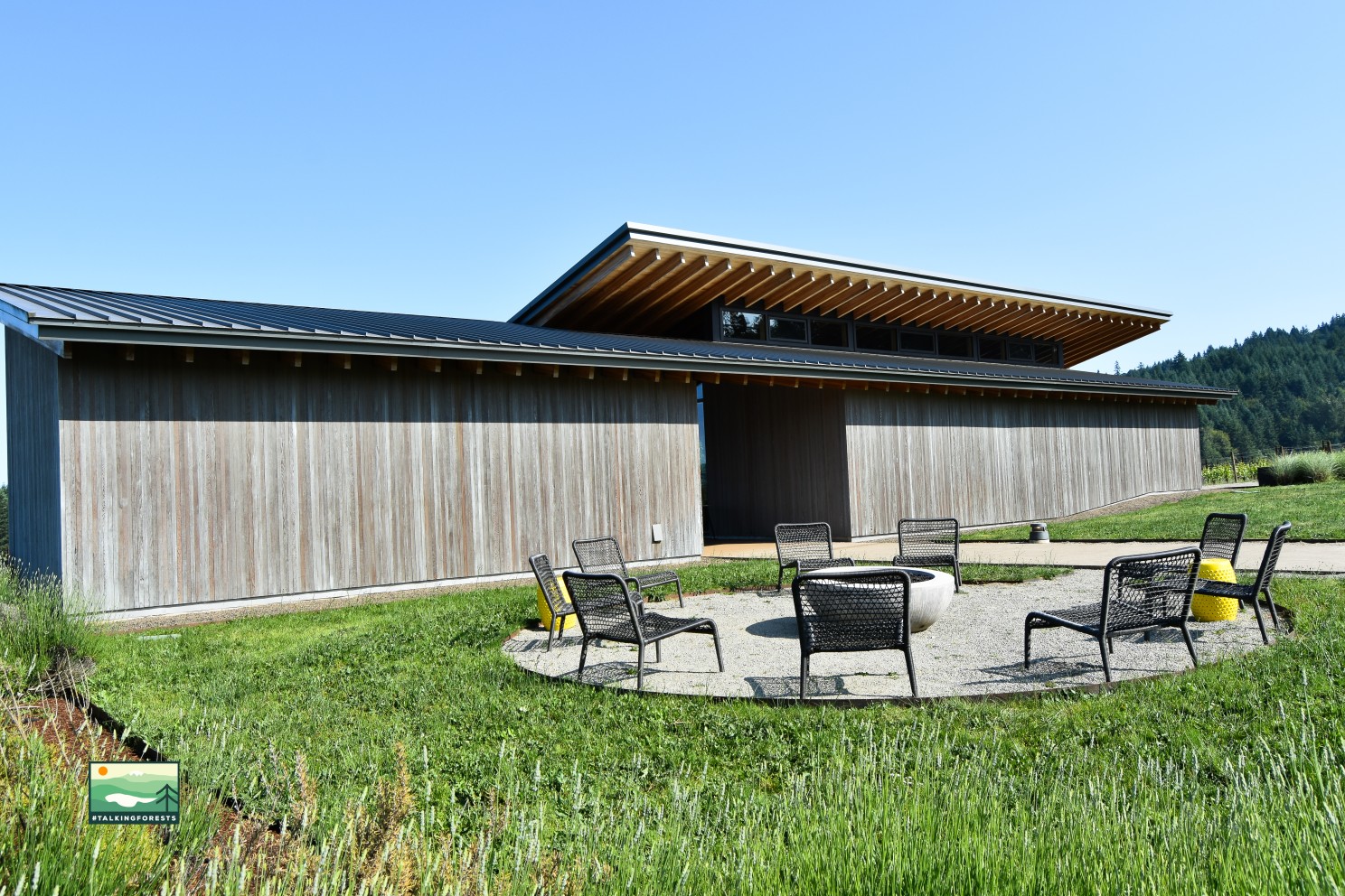  From the gravel parking lot, looking at the outside of the L'Angolo Estate Winery. The exterior is clad in durable Western red cedar siding. A set of patio chairs and a table is outside waiting for guests out front. 
