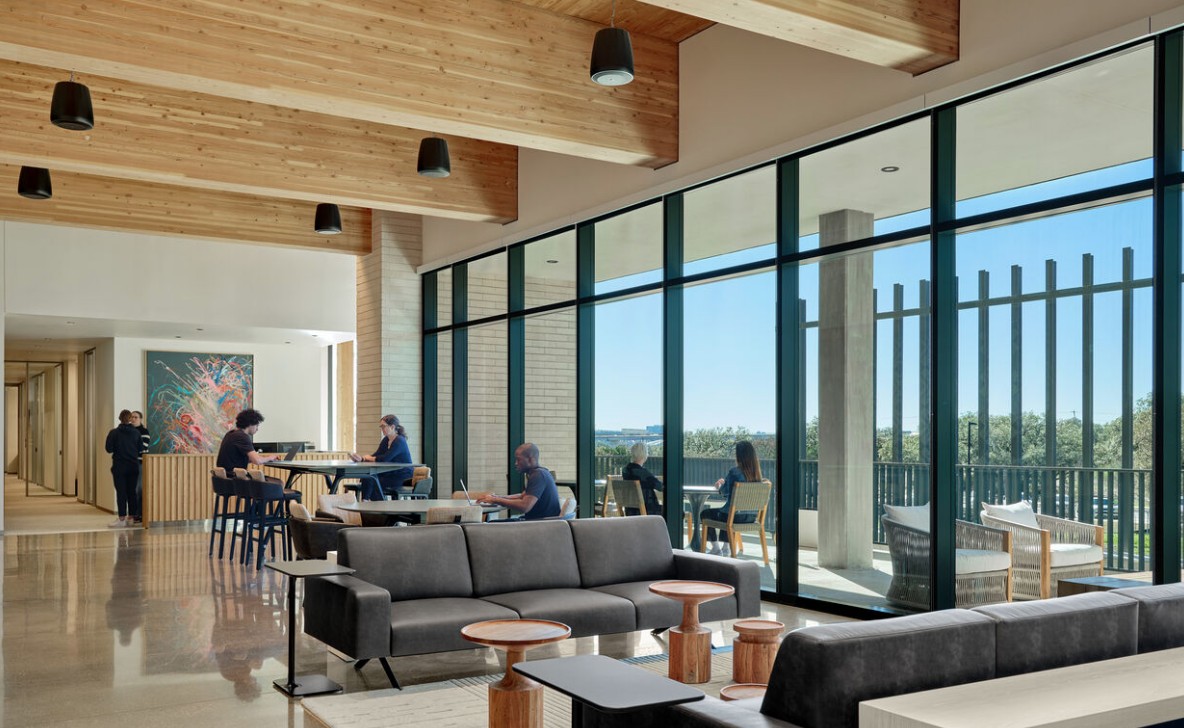 Exposed mass timber beams in a lobby with tables and people sitting around them. Comfortable furniture and side tables are near. 