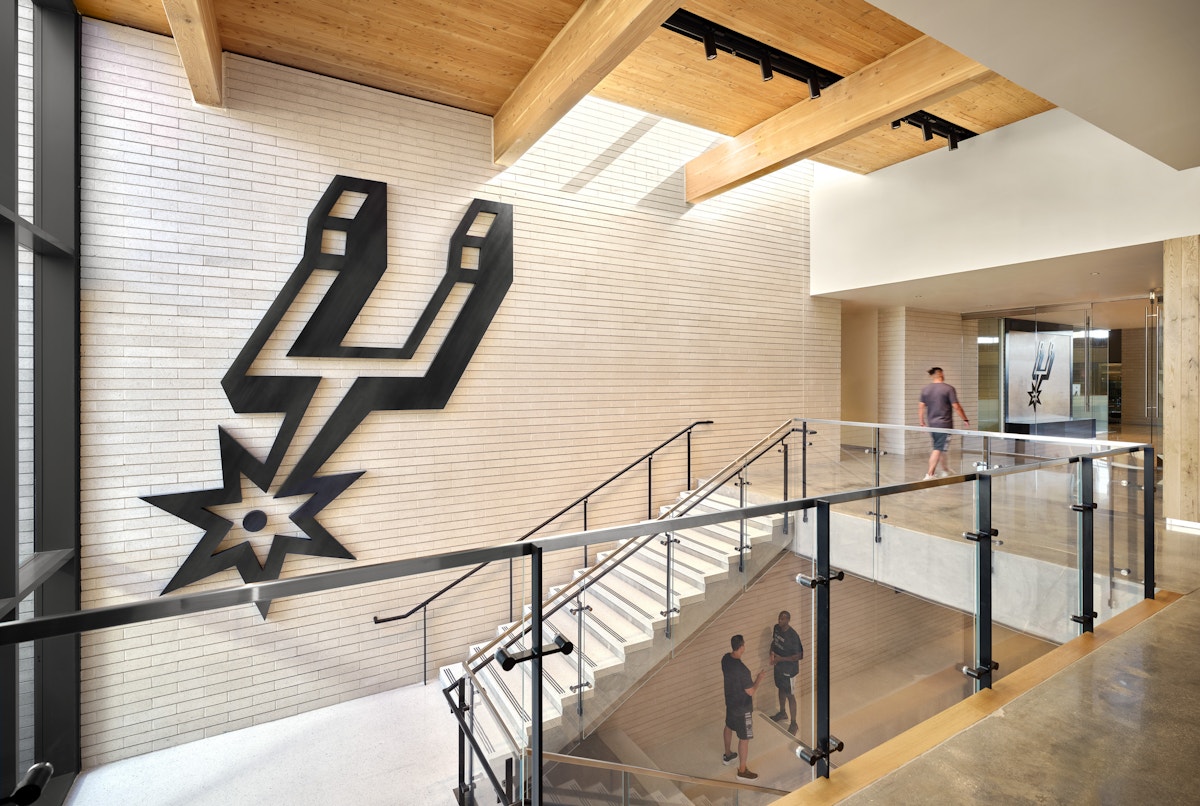 A wall with a SPURS LOGO on it. Exposed Mass Timber on the ceiling. A staircase with a glass wall showing multiple floors. 