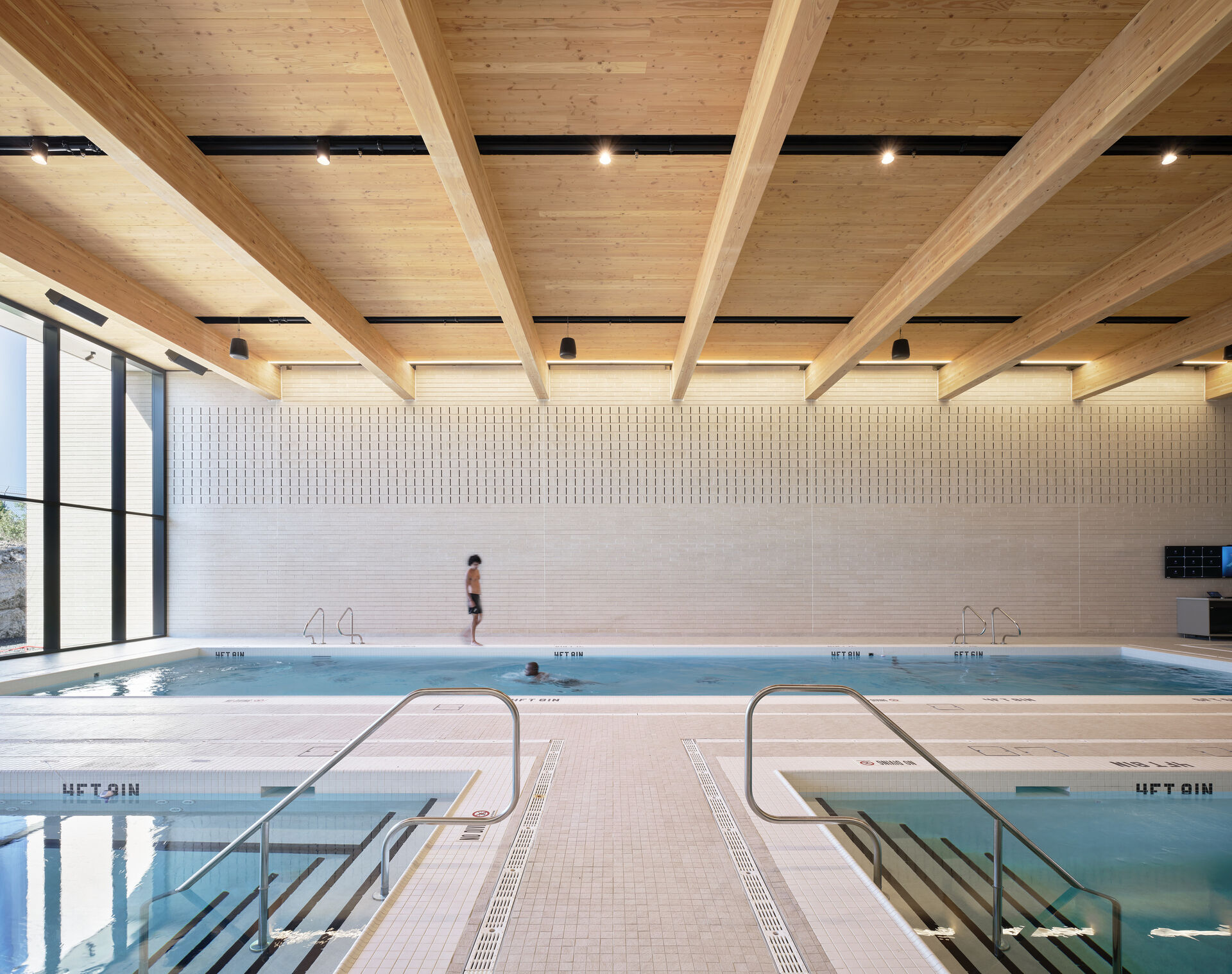 A interior picture of a swimming pool towards the back wall. Floor to celing windows are on the left. The exposed mass timber ceiling is lighted. 