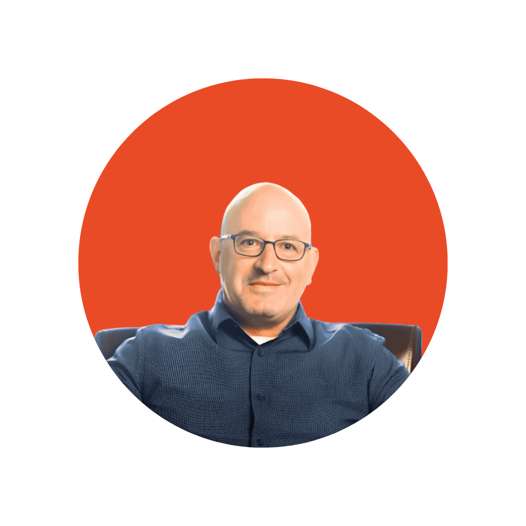 A bald man with rectangle glasses and a dark blue button down sits in a chair.