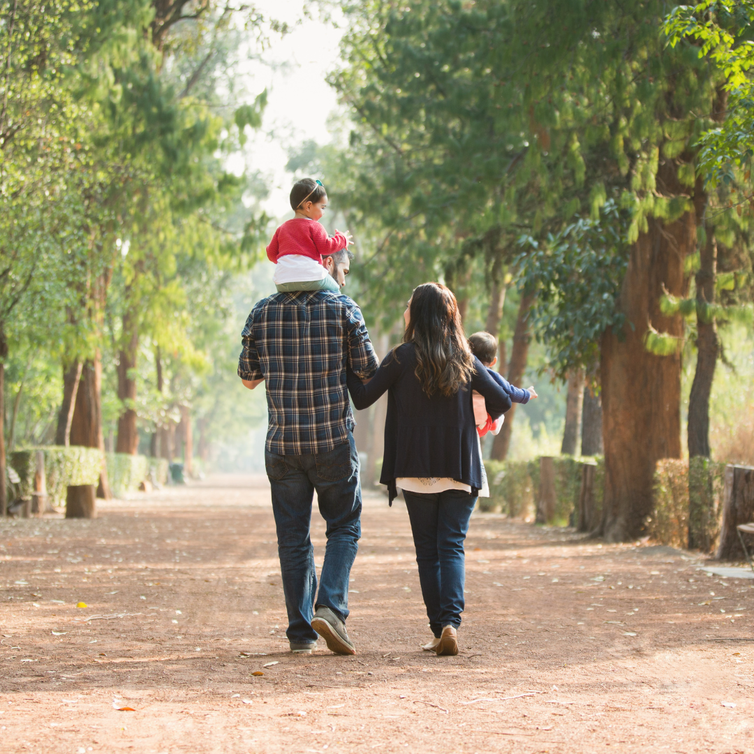 A couple walks hand in hand with their young daughter along a sunlit park path, surrounded by greenery and soft afternoon light.