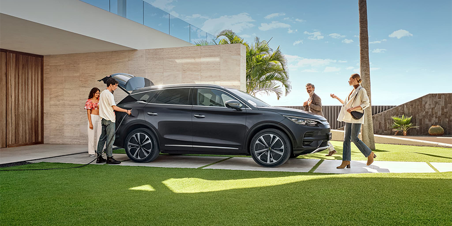 Group of people gathered around a charging BYD SUV in a modern home driveway, showcasing lifestyle benefits of electric vehicle ownership