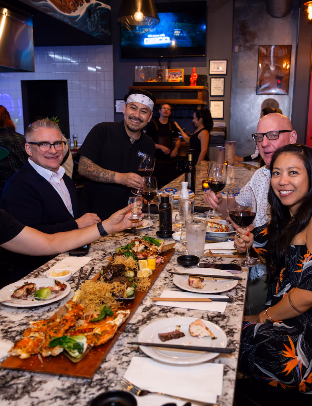 Group of five people at a restaurant table enjoying seafood dishes and wine, smiling at the camera.