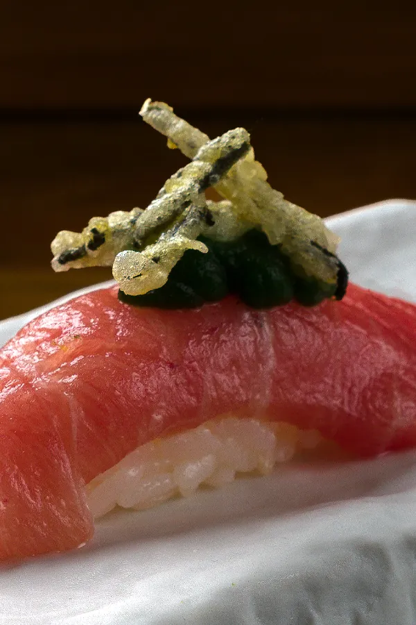 Nigiri de sushi con una rebanada de atún rosado, arroz y un topping de tempura crujiente y pasta verde.