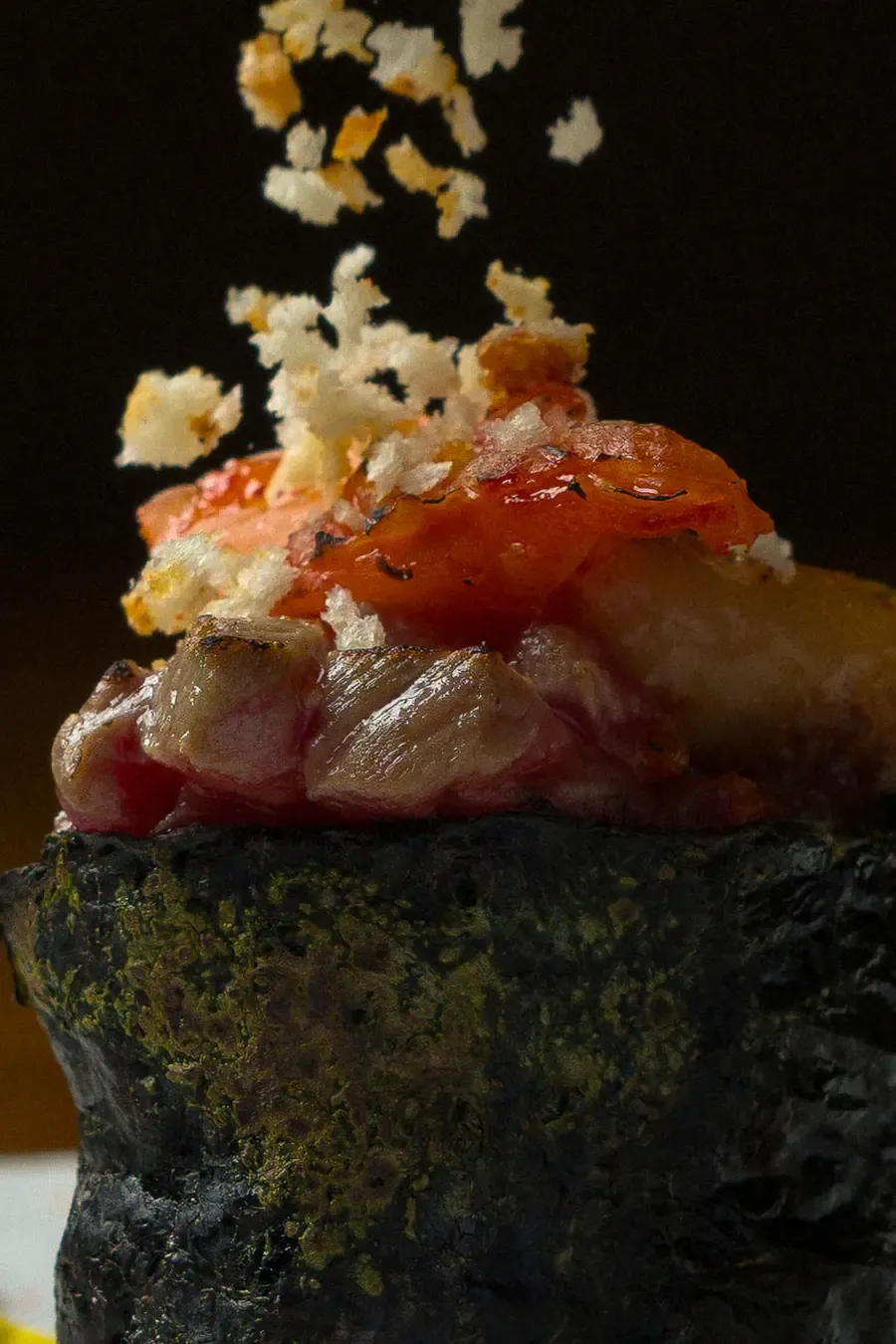 Platillo gourmet con base negra, carne asada, rodajas de tomate y migas crujientes cayendo desde arriba.