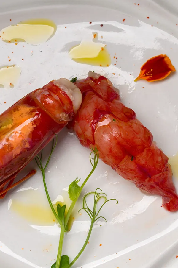 Dos langostinos crudos con piel roja brillosa acompañados de un tallo verde y gotas de aceite en un plato blanco.