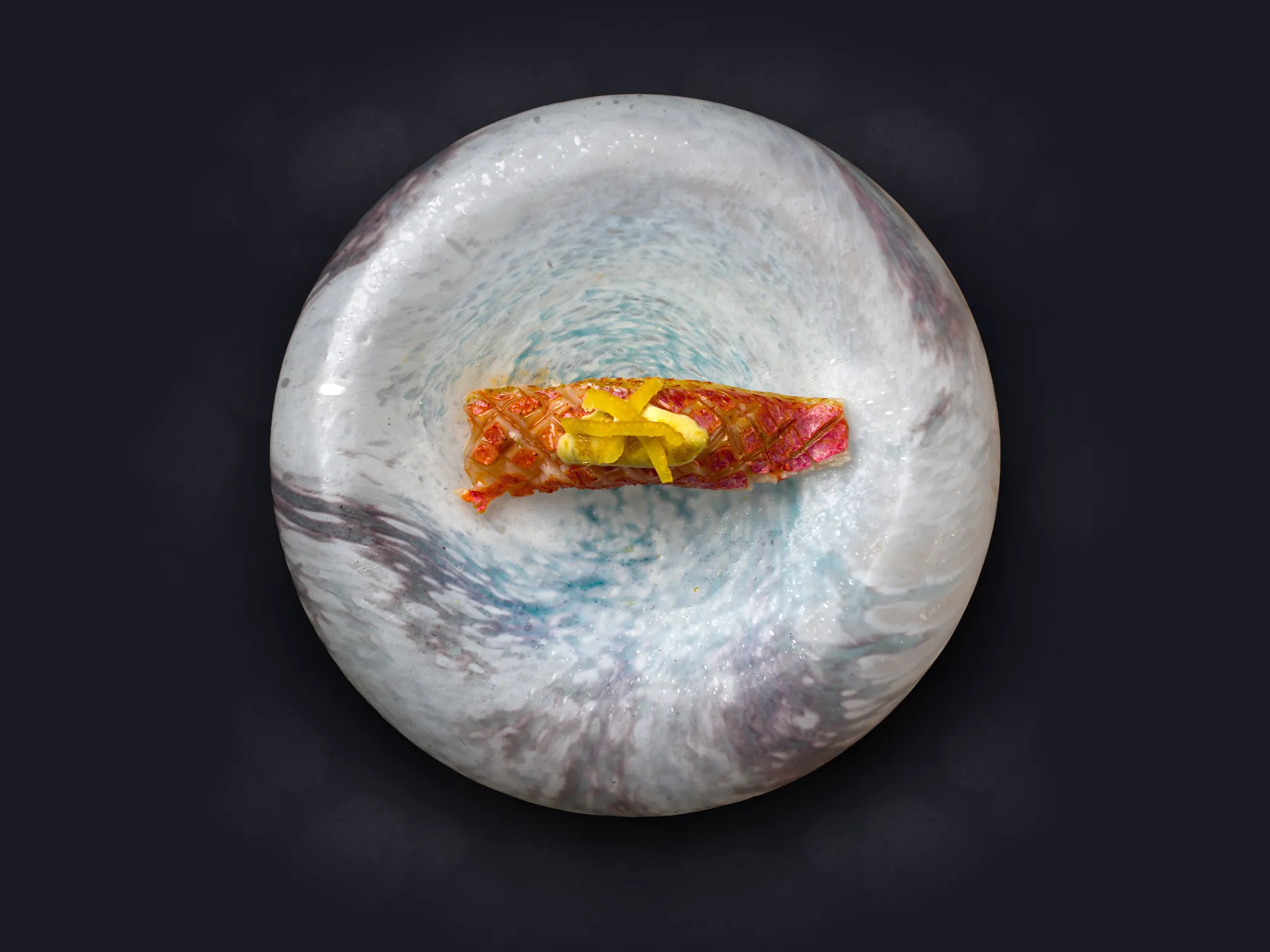 Pieza de sushi con crujiente encima y cintas de pepino servida en un plato redondo con tonos grises y azulados.