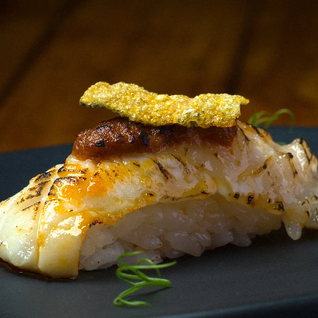 Nigiri de pescado blanco a la parrilla con salsa y crujiente dorado, servido sobre arroz al vapor.