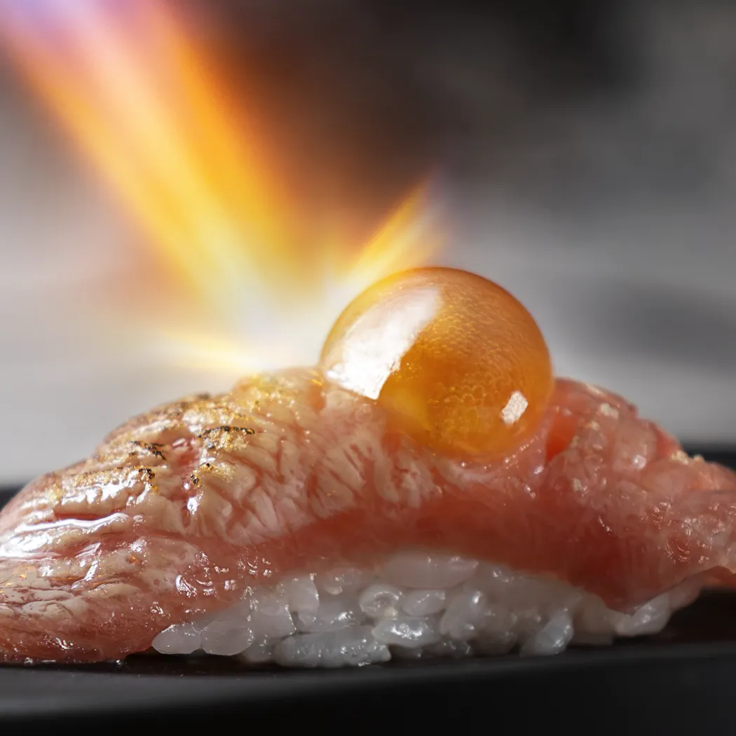Nigiri de sushi con trozo de pescado ligeramente flameado y una esfera translúcida encima, sobre arroz blanco.