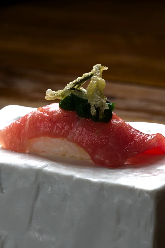 Nigiri de sushi con pescado rojo, hojas verdes y tempura encima, servido sobre una superficie blanca.