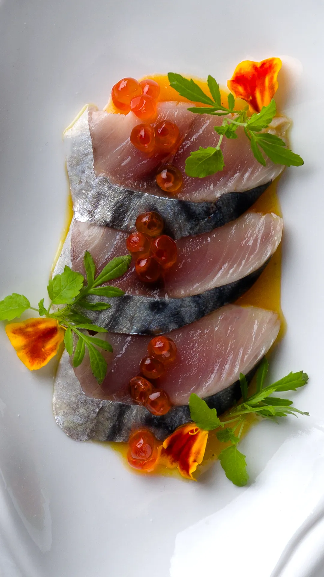 Tres filetes de pescado crudo con piel plateada, decorados con huevos de pescado naranjas, hojas verdes y flores comestibles, servidos en un plato blanco.