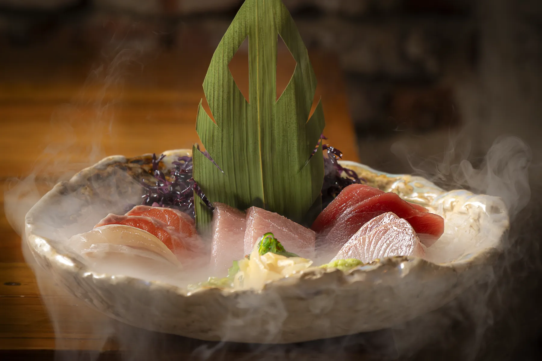 Plato de sashimi con varias piezas de pescado fresco y una hoja decorativa, servido con vapor alrededor.