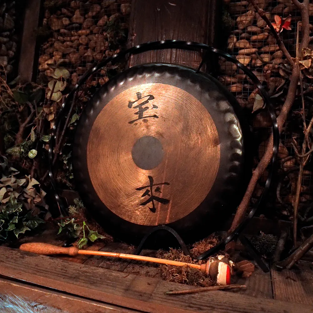 Gong de metal con caracteres asiáticos, colocado sobre una base de madera con un mazo de mango largo al frente.