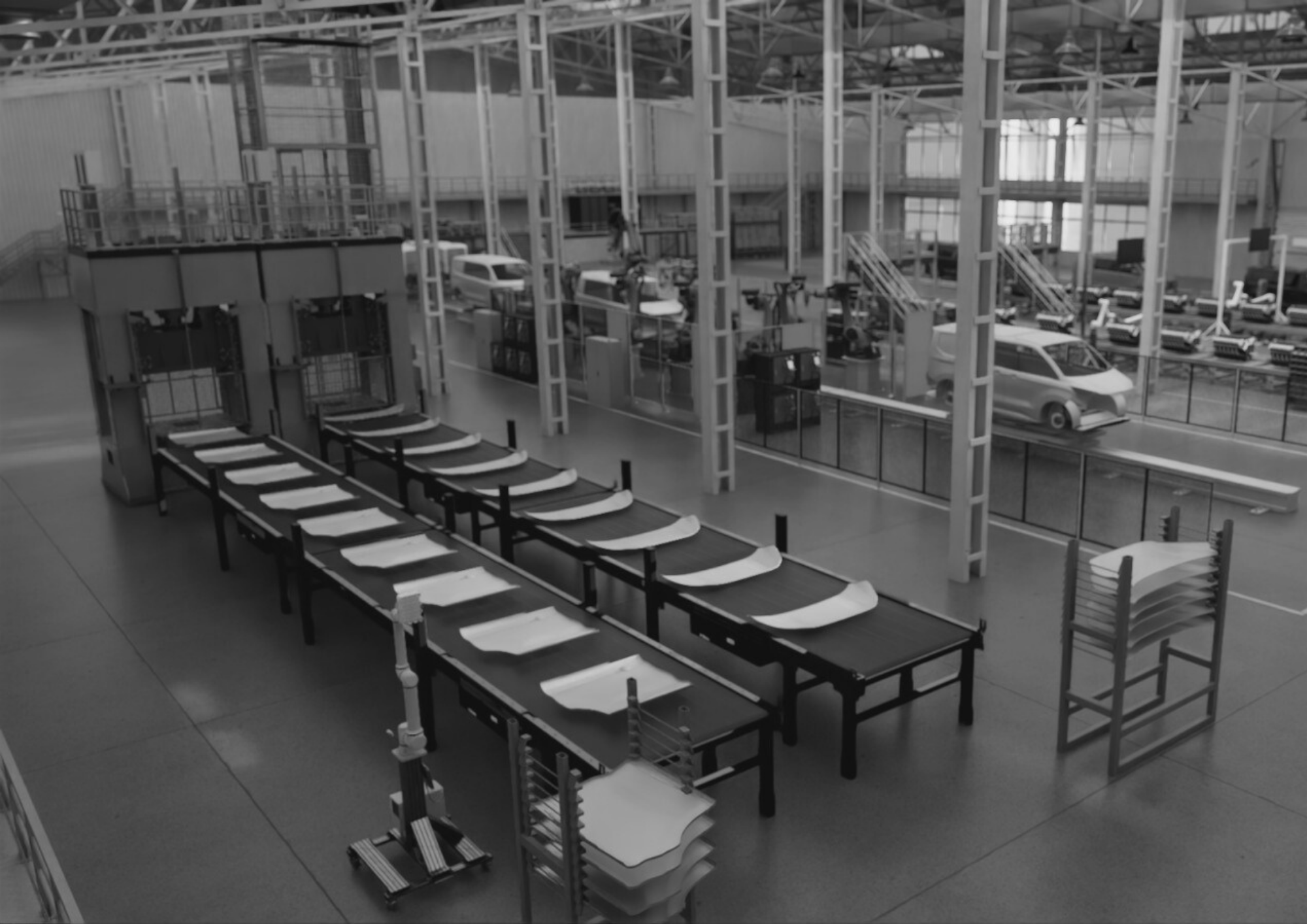 Black and white image of an automotive factory interior showing a series of conveyor tables lined with large stamped metal panels, likely body parts. In the background, industrial press machines and parked vehicles are visible, along with overhead support structures and equipment, indicating a stamping or pre-assembly area.