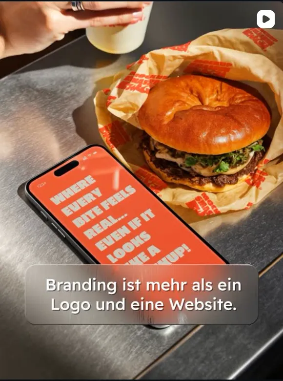 Hand hält ein Getränk über einem Hamburger in Papierverpackung auf einem Metalltisch, neben einem Smartphone mit Text auf dem Bildschirm.