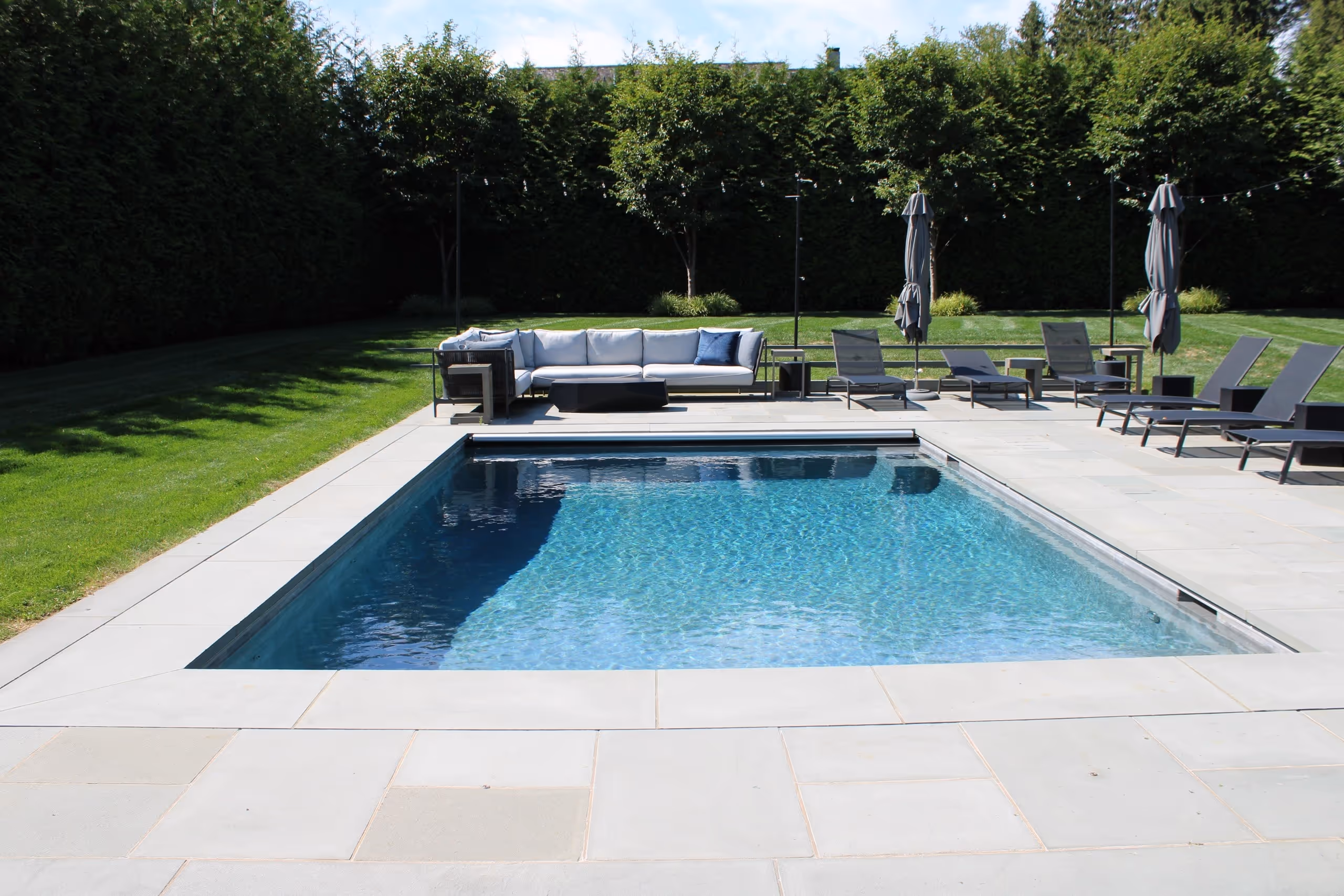 Modern backyard pool with elegant lounge seating, gray umbrellas, and manicured lawn in Greenwich, CT by New Canaan Pool Service