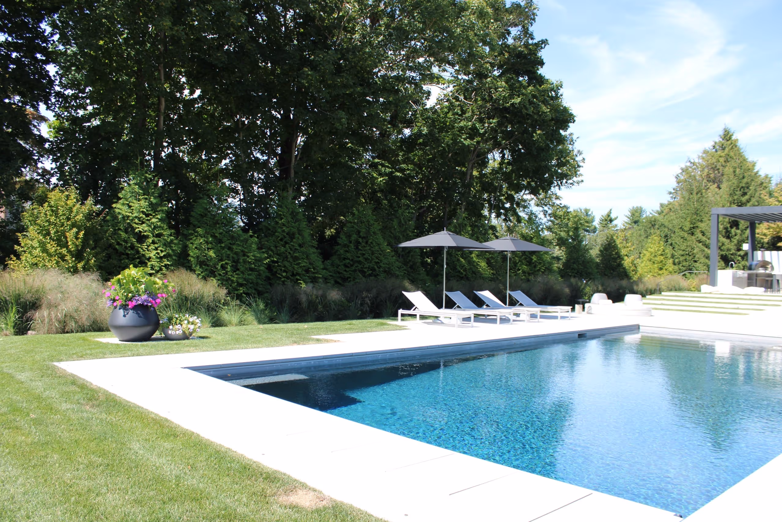 Modern inground pool with sleek lounge chairs, black umbrellas, and outdoor kitchen surrounded by lush greenery in Greenwich, CT by New Canaan Pool Service