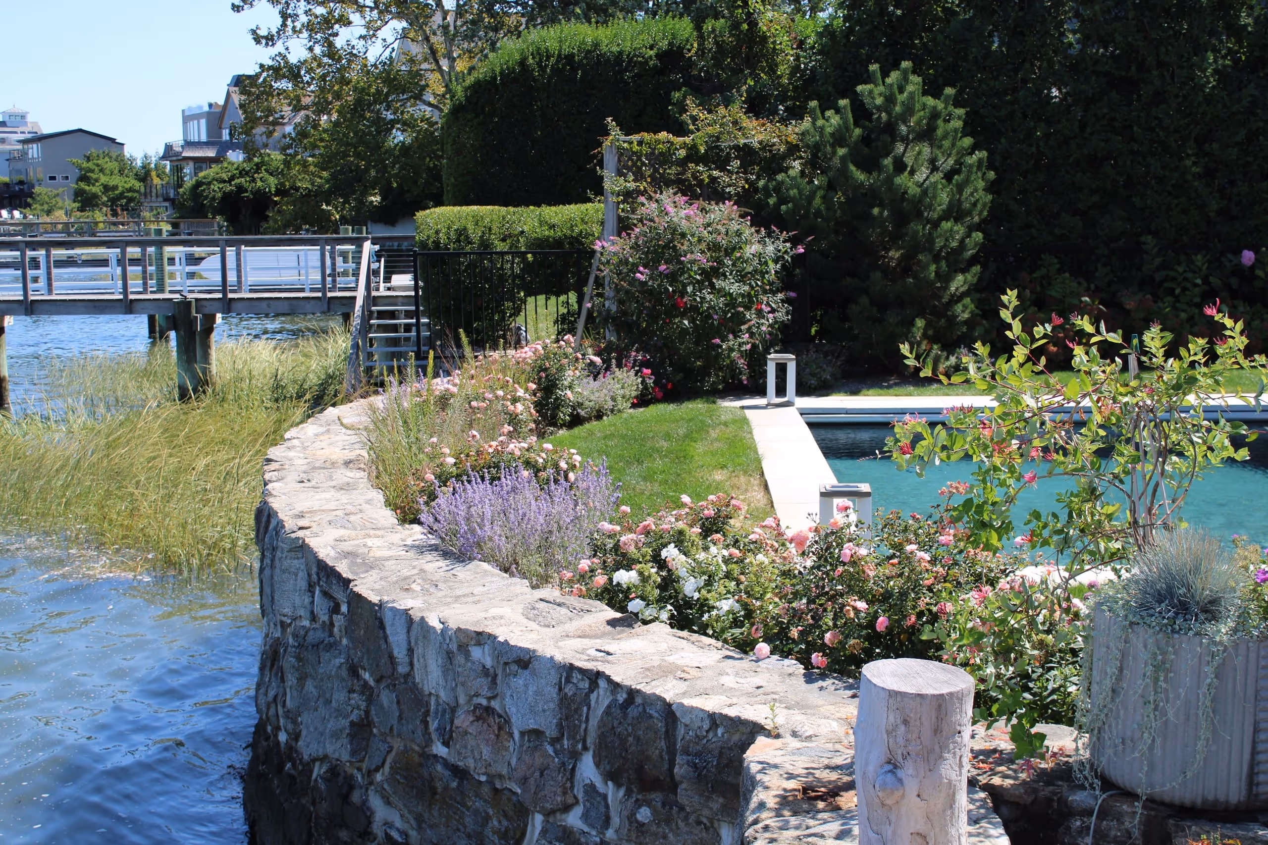 Coastal inground pool with stone retaining wall, colorful garden landscaping, and waterfront views in Darien, CT by New Canaan Pool Service