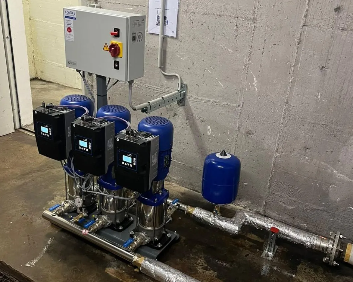 Triplex water pump system with blue motors, digital controllers, and control panel mounted on a wall in an industrial setting.