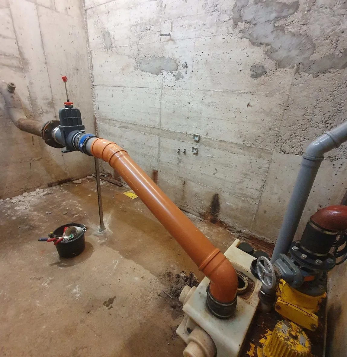 Industrial room with large orange and gray pipes connected to a yellow machine against a concrete wall.