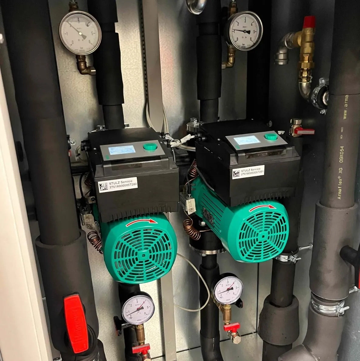 Two green and black STULZ Service pumps with pressure gauges and insulated pipes in a mechanical room.