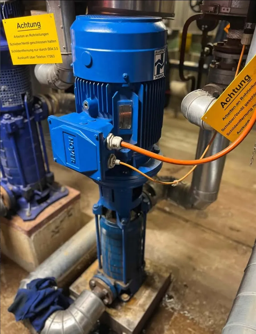 Blue industrial water pump installed with attached orange electrical cables and warning signs in German.