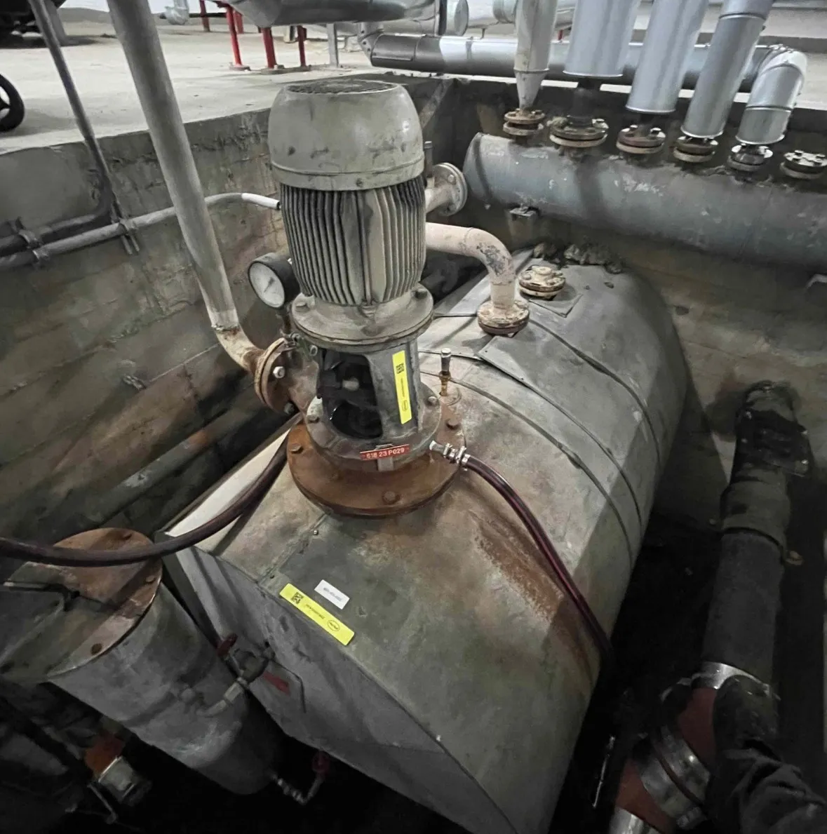 Industrial metal pump with pipes and pressure gauge installed on a cylindrical tank in a concrete basin.