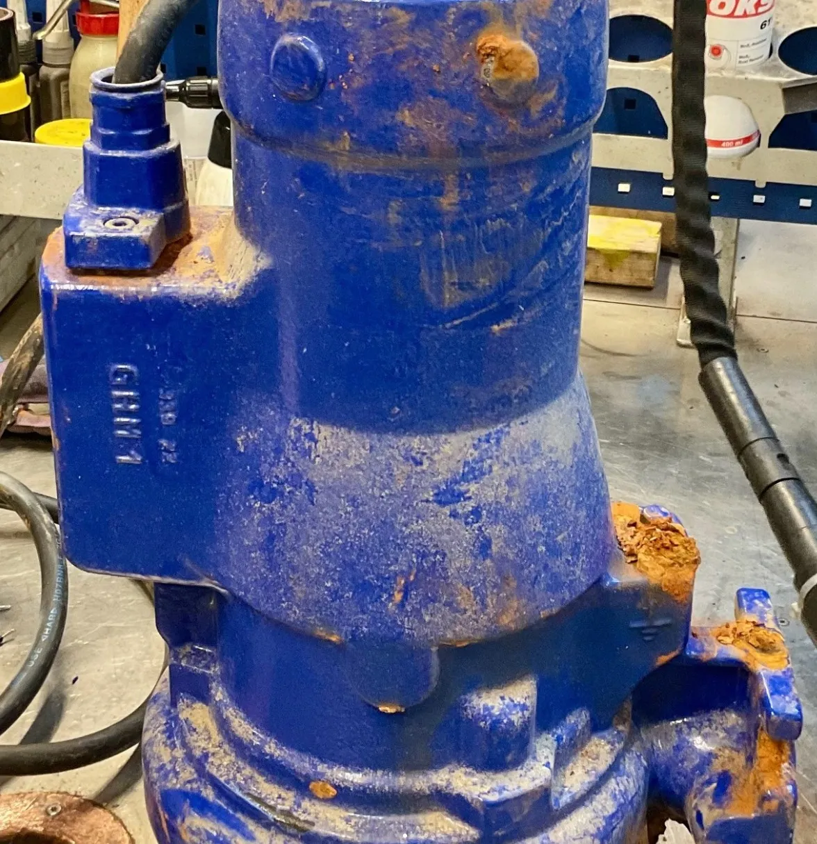 Rusty blue industrial machine part with cables and hoses on a workshop floor.