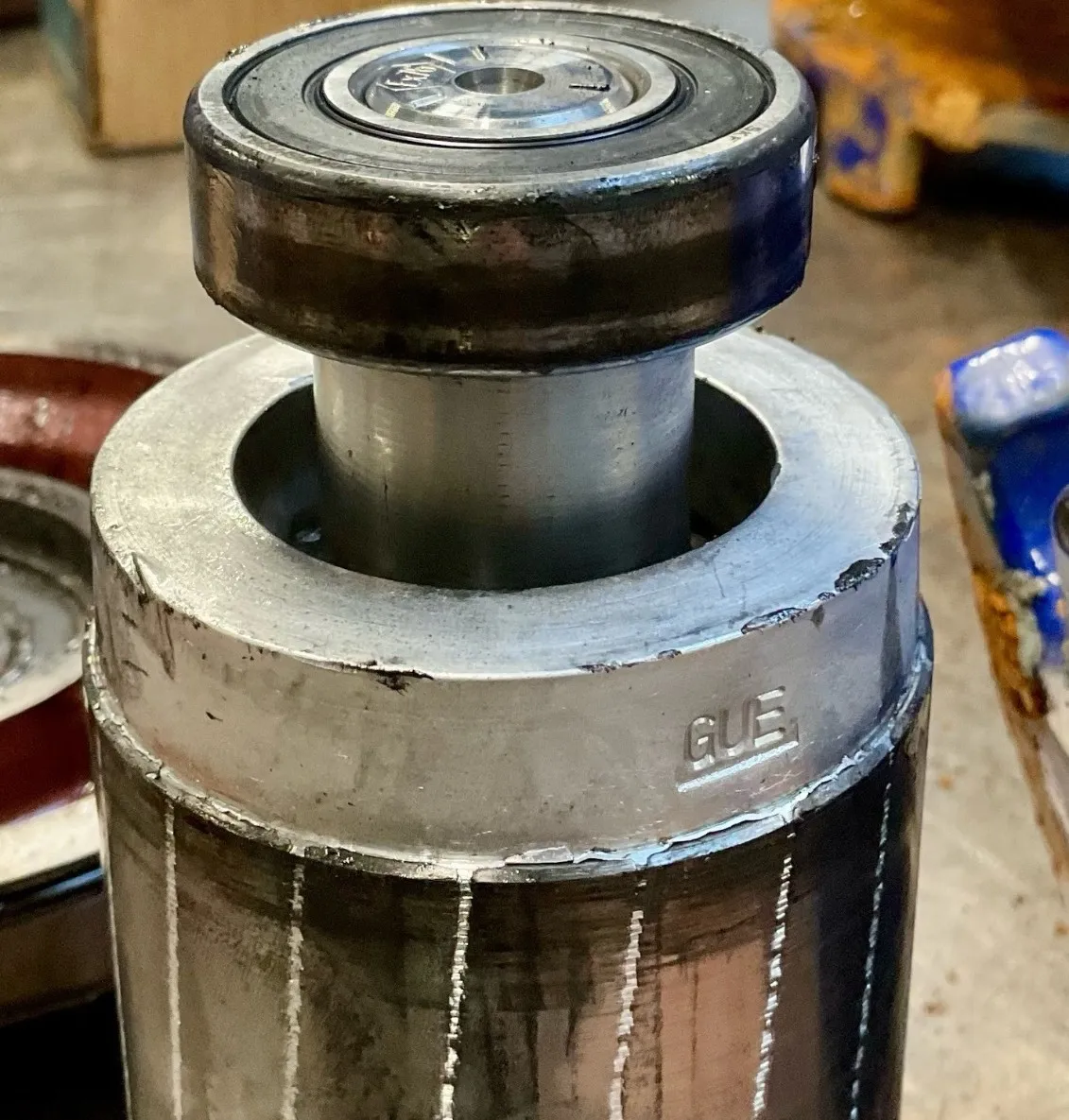 Close-up of a metallic industrial component with visible welding marks and the letters GUE stamped on the top section.