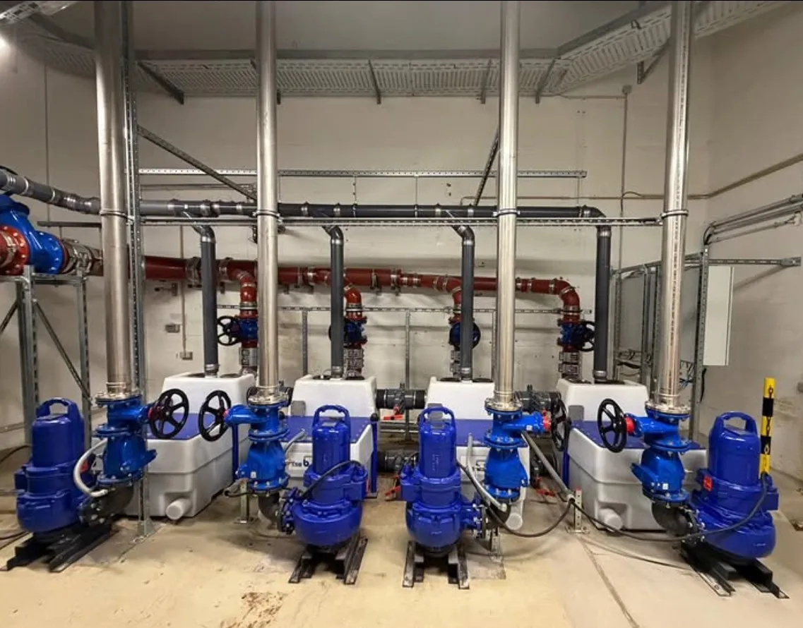 Industrial water pump system with four blue pumps connected to vertical metal pipes and white tanks in a utility room.