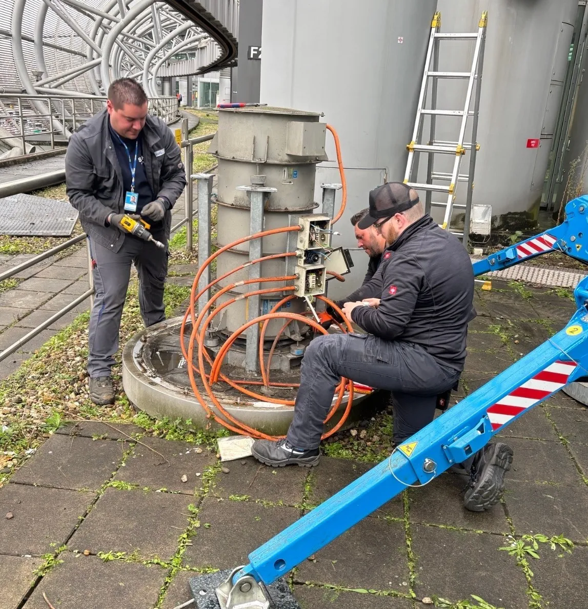 Three men working on an industrial machine with orange cables connected to it, one using a drill and two handling the cables near an electrical box outdoors.