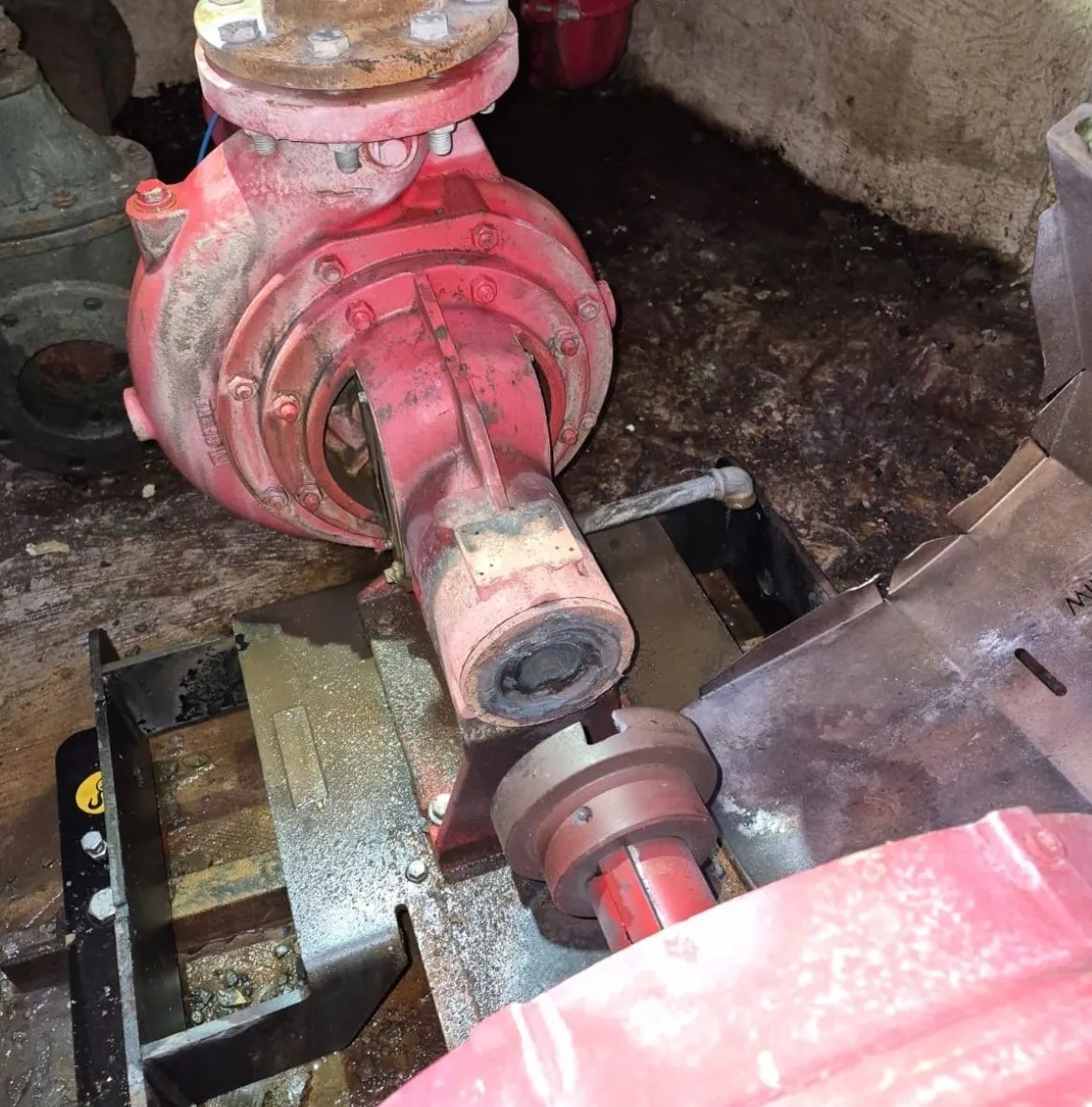 Close-up of an old, red industrial water pump with a visible coupling and metal base in a dimly lit, dirty environment.