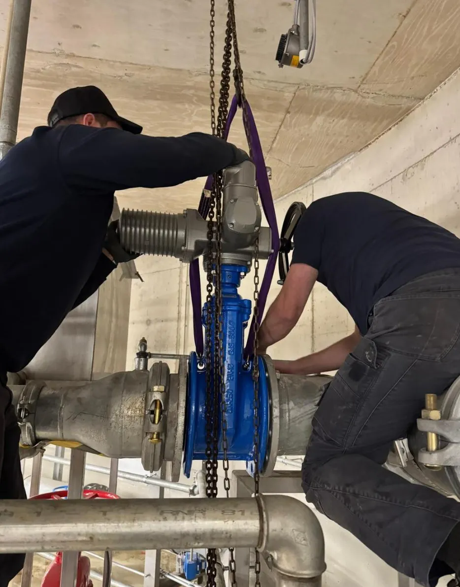 Two workers install a large blue industrial valve in a pipe system inside a concrete facility.