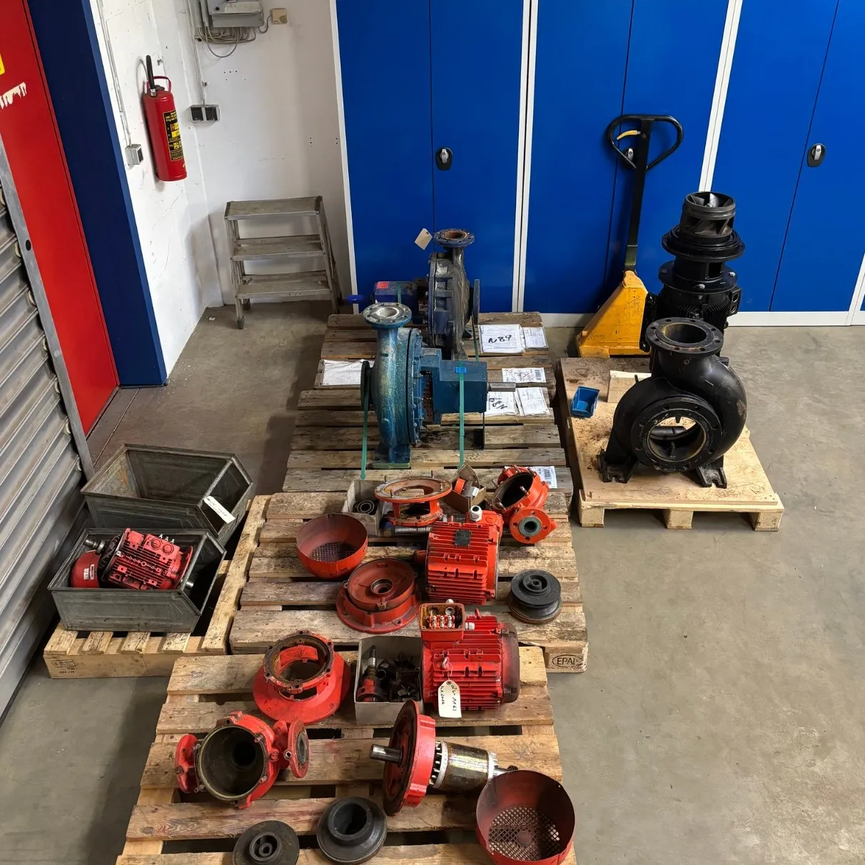 Industrial workshop floor with blue cabinets, pallets holding various metal pump parts and motors, a yellow pallet jack, and a fire extinguisher on the wall.