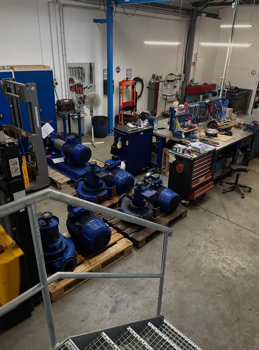 Industrial workshop with several large blue electric motors on pallets, tool benches with organized tools, and a staircase leading down to the floor.