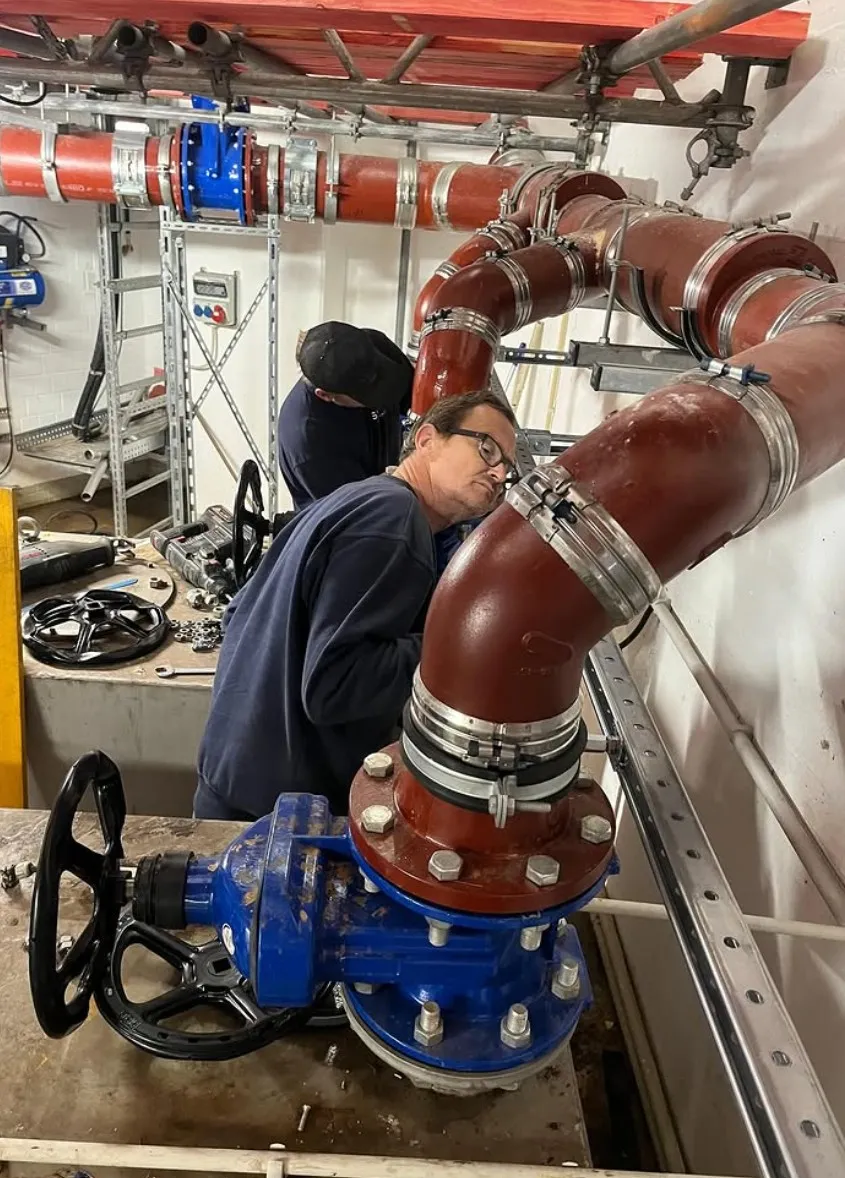 Two workers inspecting and working on large red and blue industrial pipes with valves indoors.