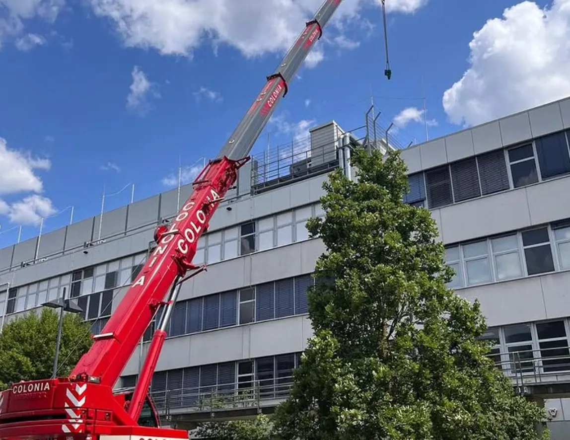 Red crane labeled 'COLONIA' lifting near a modern multi-story building with large windows under a partly cloudy sky.