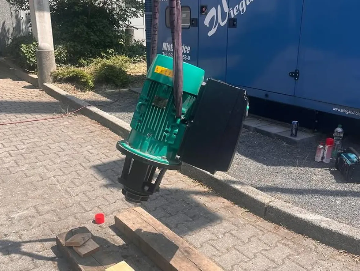 Green industrial motor suspended by purple straps over wooden planks on a paved surface next to a blue container.