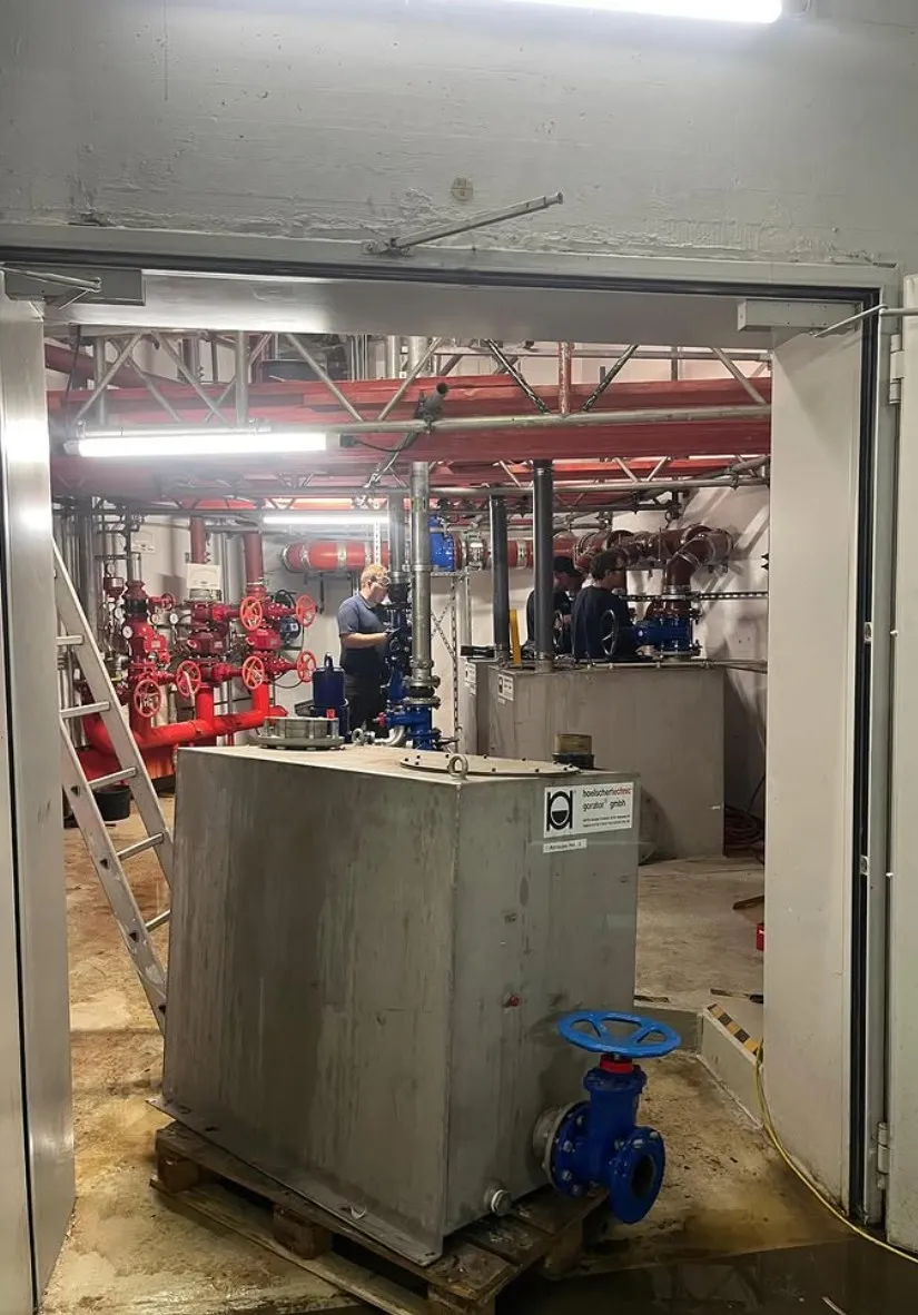 Industrial room with large metal tanks, red and blue valves, pipes, and two workers inspecting equipment.