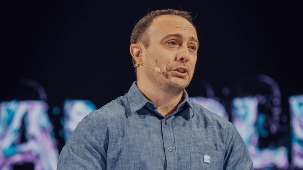 Man in a blue button-up shirt speaking with a headset microphone against a dark background.
